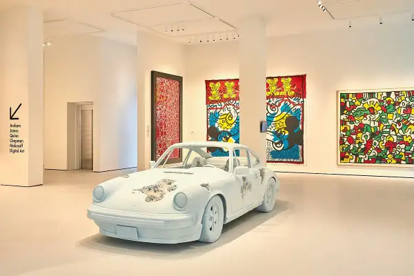 A white Porsche, artfully distressed, sits in a gallery. Vibrant Keith Haring-esque artworks adorn the walls, making for excellent Moco Museum reviews.