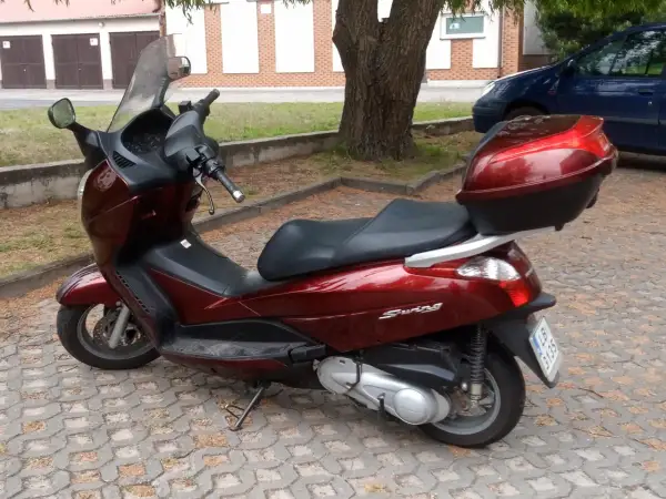 Ciemnoczerwony skuter Honda Swing 125 z czarnym siedzeniem i kufrem, zaparkowany na kostce brukowej.