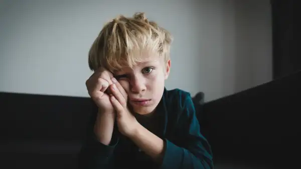 Chłopiec z blond włosami, zamyślony, opiera głowę na dłoniach. Jego spojrzenie sugeruje trudności, być może związane z zespołem Aspergera.