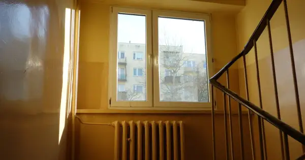 Słoneczne promienie padają na błyszczącą ścianę w klatce schodowej. Widać okno z widokiem na blok i kaloryfer.