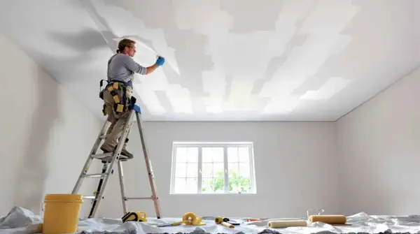 Un peintre sur une échelle applique une toile de rénovation plafond. Des rouleaux et un seau sont prêts.