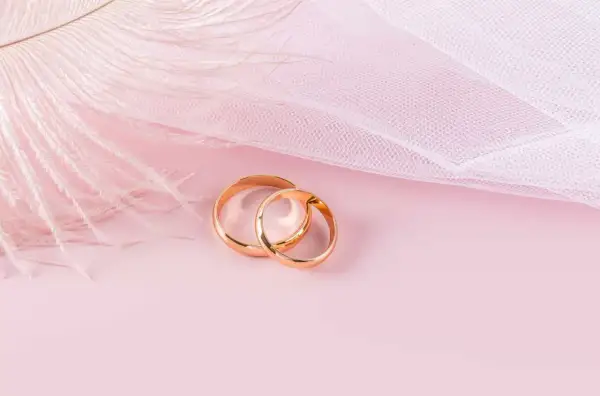 Dos anillos de oro rosa entrelazados, sobre un fondo rosa pálido con una pluma blanca y tela delicada.
