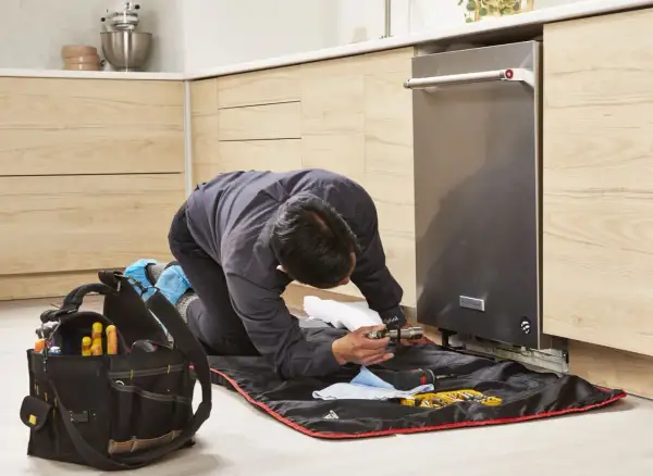 A technician kneels to replace a dishwasher, surrounded by tools and a protective mat.