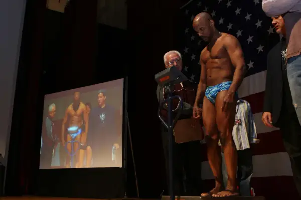 A muscular man in blue patterned shorts stands on a scale, preparing for his junior bantamweight weight check. An official looks on.