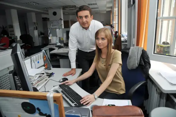 Ewa Bugała i jej mąż pracują razem w biurze. Ona siedzi przy komputerze, on stoi obok.