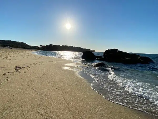 La **playa de Tulla** bajo un sol radiante. Huellas en la arena dorada y el mar azul acarician las rocas oscuras.