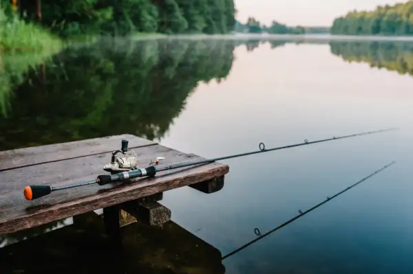 Wędka i kołowrotek leżą na drewnianym pomoście, gotowe do wędkowania na Mazurach. Spokojna tafla jeziora odbija zalesione brzegi.
