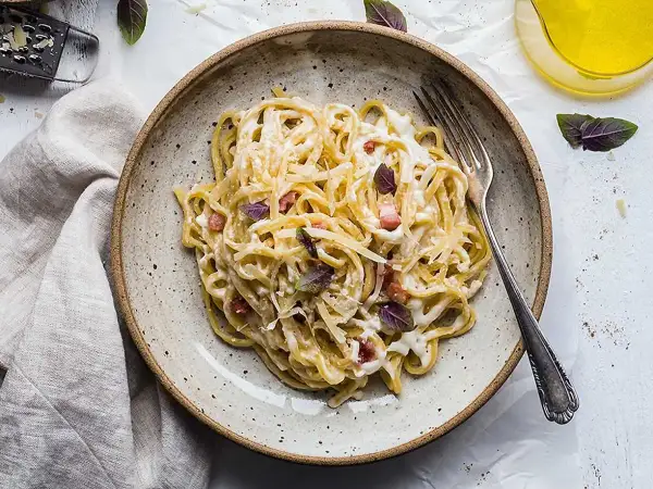 En tallrik med krämig, klassisk carbonara, toppad med basilika och parmesan. En gaffel ligger redo att avnjutas.