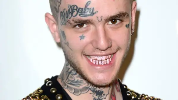 Uśmiechnięty Lil Peep z tatuażami na twarzy i szyi, błyszczącymi różowymi zębami. Pytanie czy lil peep żyje, pozostaje w sercach fanów.
