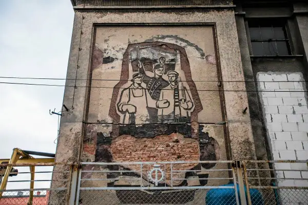 Zniszczony mural w stylu socrealizmu przedstawiający trzech robotników z karabinami.
