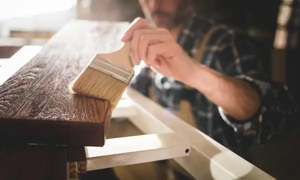 Un falegname applica una finitura protettiva su un pezzo di legno. Dopo l'impregnante, cosa si mette? Una vernice trasparente o una cera per esaltare la venatura.