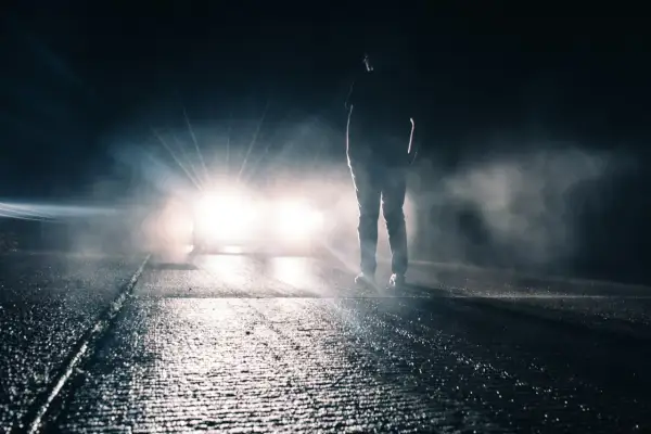 Silhouette d'une personne sur une route mouillée, éclairée par un appel de phare éblouissant dans la nuit brumeuse.