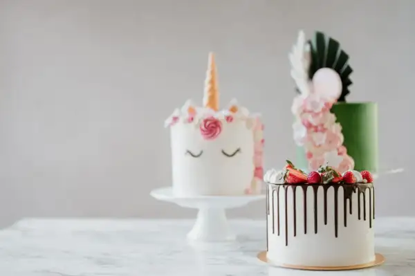 En förtrollande rosa tårta i enhörningsstil, bredvid en grön tårta med blommor och en vit tårta med chokladdryp och bär.