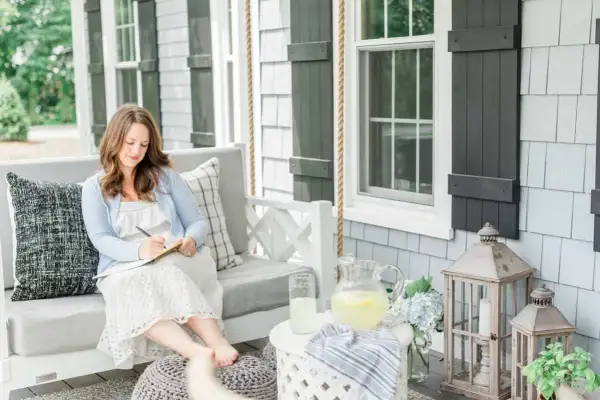 A woman embodies the coastal grandma aesthetic, writing in a notebook on a porch swing, with lemonade and hydrangeas nearby.