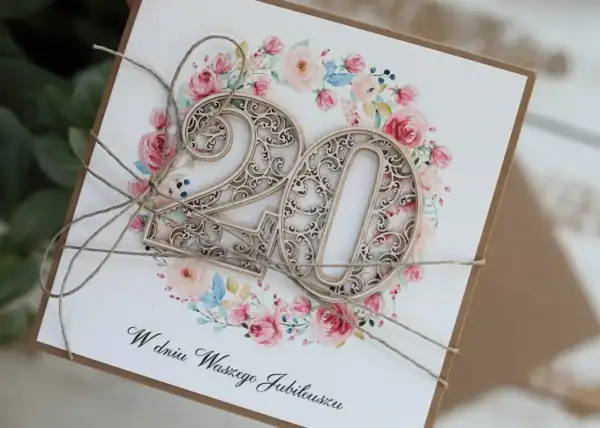 Elegancka kartka z napisem "W dniu Waszego Jubileuszu" i ażurowymi cyframi "20" na tle kwiatowej girlandy. Idealna na 20 rocznicę ślubu życzenia.