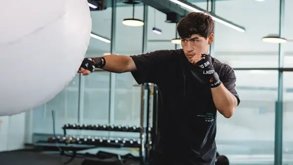 A young man practices Russian boxing, throwing a punch at a large white training bag in a modern gym.