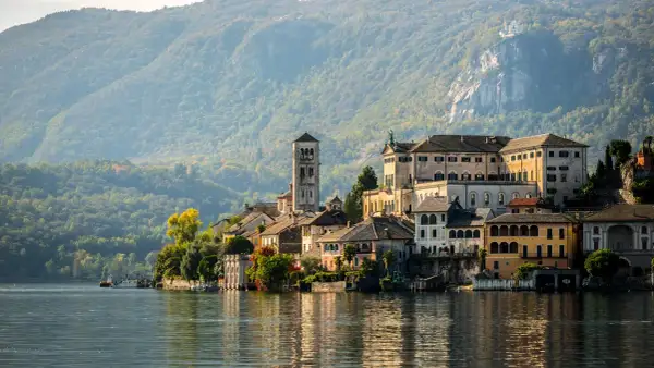 A picturesque Italian village with colorful buildings and a tall bell tower sits on the edge of a calm lake, with tree-covered mountains in the background.
