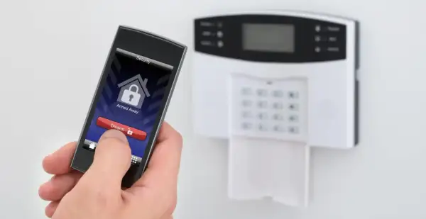 Dłoń trzyma smartfon z aplikacją do ochrony mieszkania w bloku. Na ekranie widnieje przycisk "Disarm". Obok panel alarmowy.