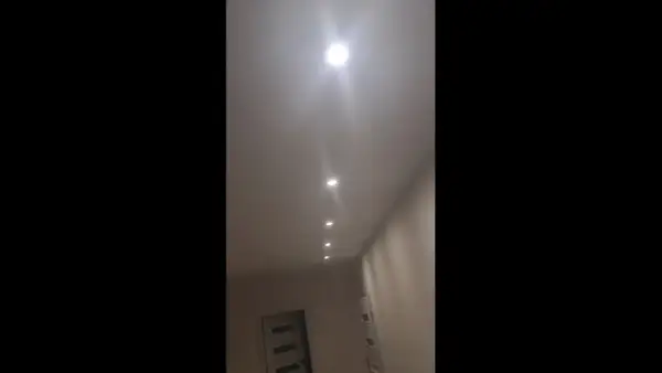 W korytarzu domu widać rząd wbudowanych w sufit lamp. Jedna z nich, najbliżej widza, świeci najjaśniej, tworząc efekt migającego światła.