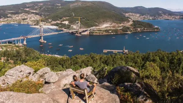 Pareja sentada en un mirador, contemplando el impresionante puente de Rande sobre el mar, con bateas de mejillones y un pueblo al fondo.