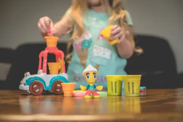 Dziecko bawi się w play doh warsztat, tworząc lodowe przysmaki. Na stole kolorowe foremki i figurka lodziarza.