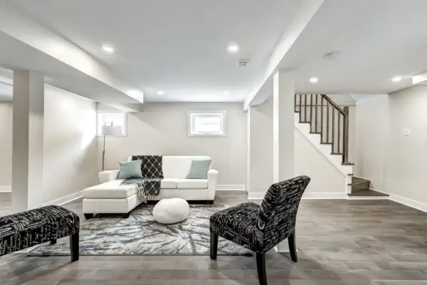 A bright, modern living area in a finished basement, featuring a white sectional, patterned rug, and a staircase leading up.