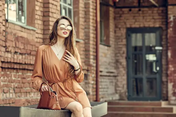 Kobieta w brązowej sukience i okularach przeciwsłonecznych, z torebką. Idealne buty do brązowej sukienki? Zobacz stylizację!