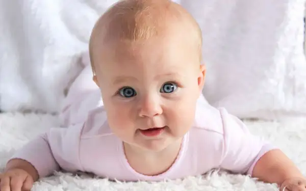 Bebé con ojos azules y cabello rubio, con un mameluco rosa. Pensando en nombres de chica ingleses como Emily o Olivia.