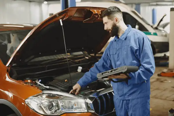 Mécanicien en bleu effectuant un diagnostic électronique sur un moteur de voiture orange. Il tient un appareil de diagnostic, essentiel pour connaître le prix d'un diagnostic voiture.