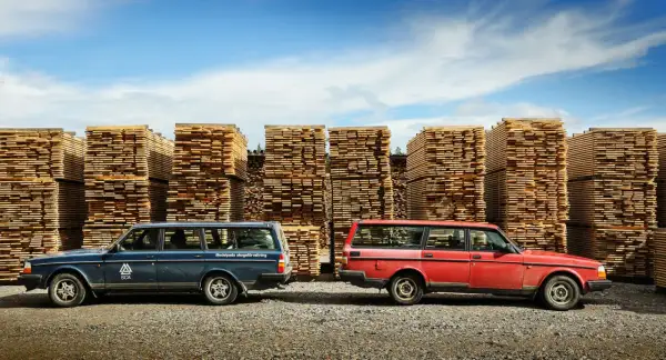 Två Volvo 240 kombi, en blå och en röd, står framför höga staplar av träplankor under en blå himmel.