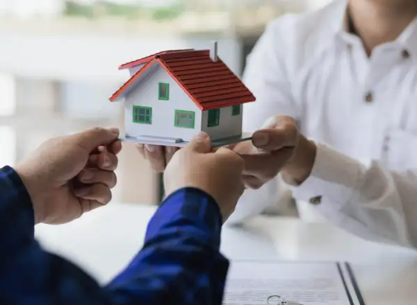 Manos intercambian una maqueta de casa, simbolizando la decisión de vender herencia.