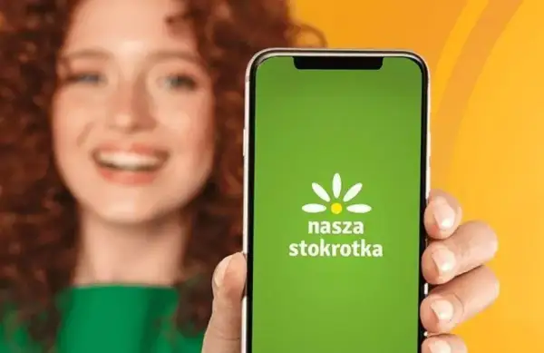 Uśmiechnięta kobieta prezentuje ekran telefonu z zieloną aplikacją "Nasza Stokrotka".