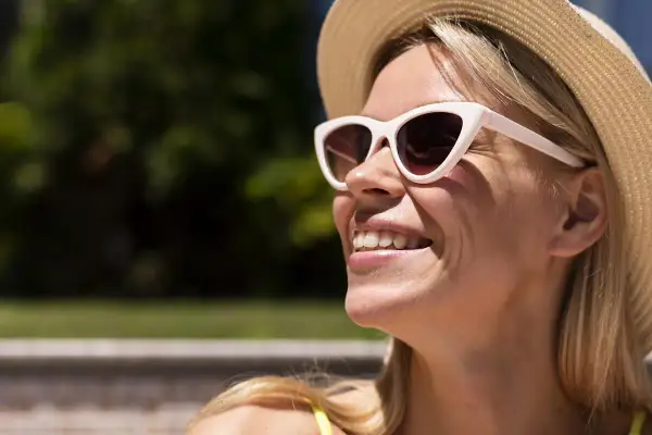 Donna sorridente con cappello di paglia e occhiali da sole, le rughe da sole che le solcano il viso.