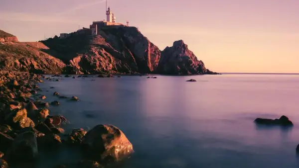 Faro en un acantilado rocoso al atardecer, con el mar tranquilo y piedras en la orilla. Parece la boca de los frailes.