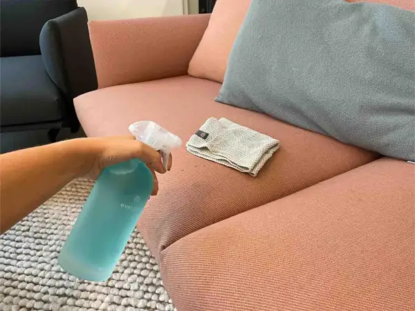 Hand sprüht Reinigungsmittel auf einen rosafarbenen Sofa-Stoff, um Wasserflecken zu entfernen. Ein Tuch liegt daneben.
