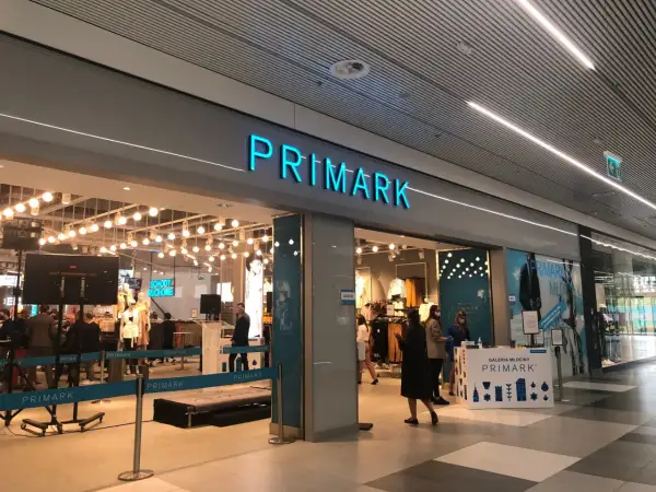Wejście do sklepu Primark. W środku widać manekiny i ubrania. Na ladzie informacyjnej znajduje się **Primark tabela rozmiarów**.