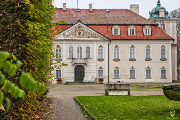Pałac w Nieborowie z czerwoną, ceramiczną dachówką i ozdobnym frontonem. Wokół jesienne liście i bujna zieleń.
