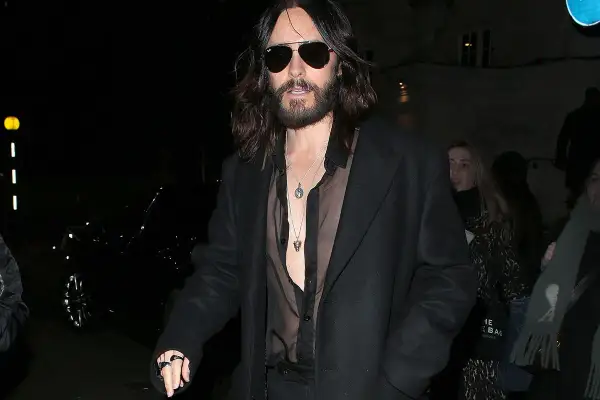 Jared Leto con un audace outfit camicia nera trasparente sotto un cappotto scuro, collane d'argento e occhiali da sole.