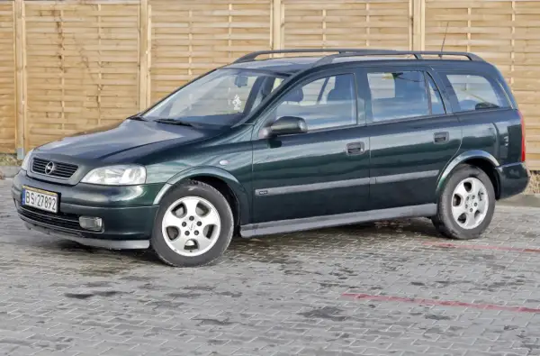 Zielony Opel Astra kombi, jeden z wielu aut w ocynku, stoi na mokrym parkingu przed drewnianym płotem.