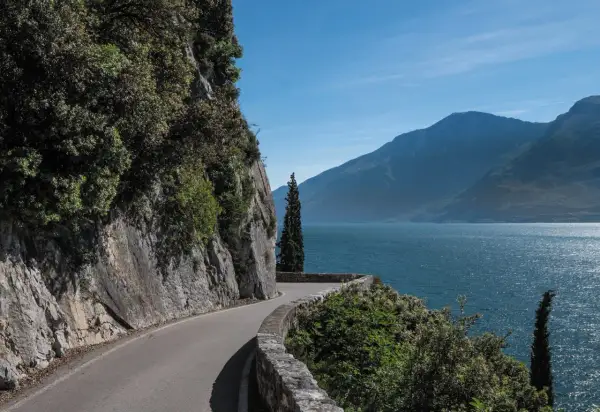 Strada panoramica lungo il lago, con rocce a sinistra e montagne sullo sfondo. La gardesana occidentale offre scorci mozzafiato.