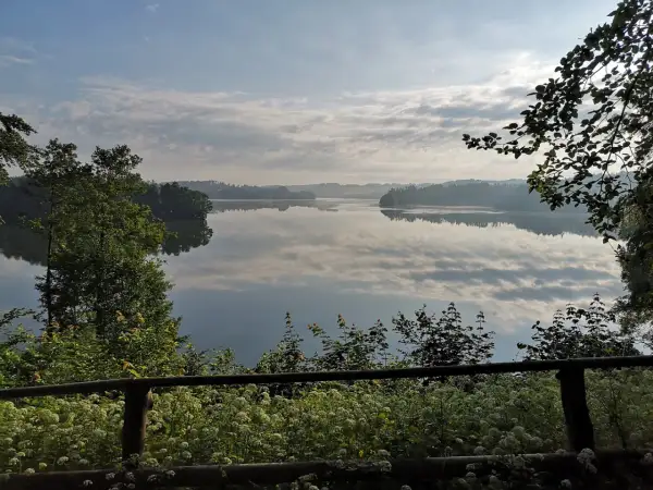 Spokojny poranek nad jeziorem w Sulęczynie. Lustrzana tafla wody odbija chmury i zalesione brzegi.