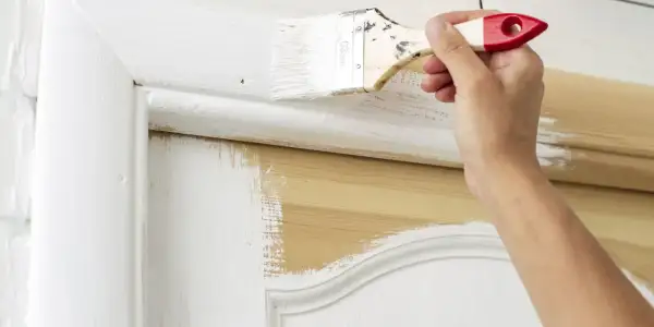 Une main applique de la peinture blanche sur une porte intérieure en bois. Le travail de peinture est en cours, révélant le bois naturel sous la couche blanche.