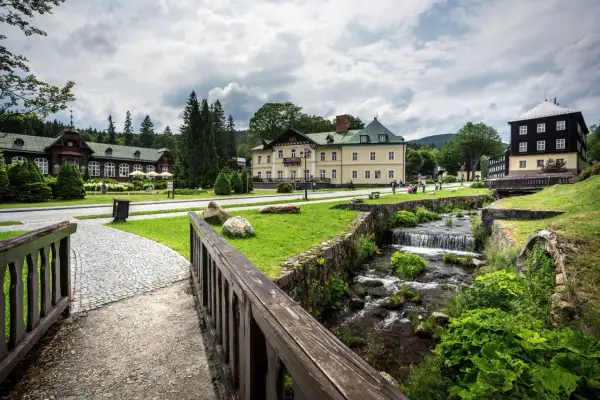 Idyllische Szene in Karlova Studanka: Gebäude im Kurort, ein Bach plätschert über Steine, umgeben von Grün.