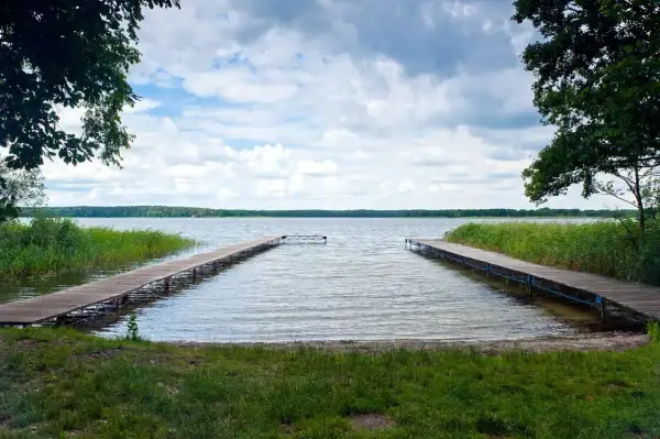 Dwa drewniane pomosty prowadzące na spokojne jezioro, otoczone zielenią. Idealne miejsce na relaks i podziwianie przyrody, jedna z wielu atrakcji w Boszkowie.