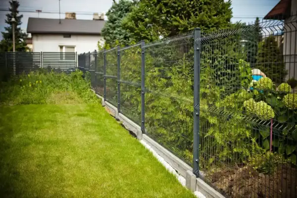 Solidne, metalowe pełne ogrodzenie od sąsiada oddziela zadbany trawnik od bujnej zieleni i domu.