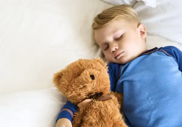 En liten pojke sover sött med sin nallebjörn. Hur många timmar ska en 10-åring sova? Sömn är viktigt för barn.