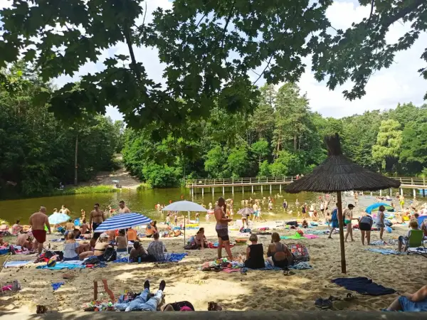 Słoneczny dzień na plaży przy zalewie w Garbatce. Ludzie odpoczywają na piasku, kąpią się w wodzie i spacerują po pomoście.