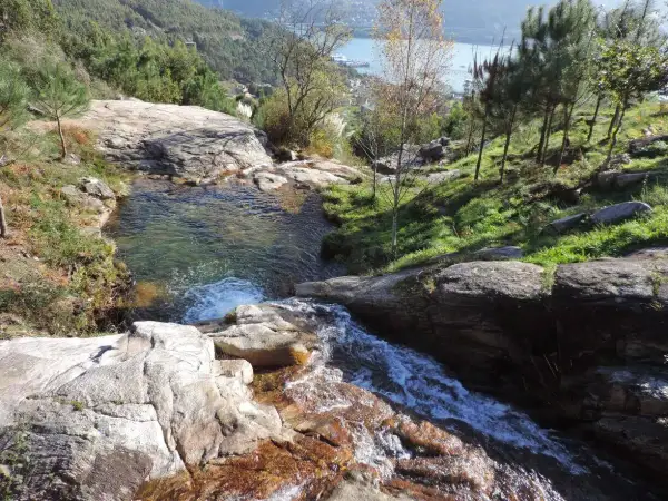 Cascada y pozas naturales en Pozas da Moura, con agua cristalina cayendo sobre rocas y vegetación exuberante. Al fondo, un estuario y colinas verdes.
