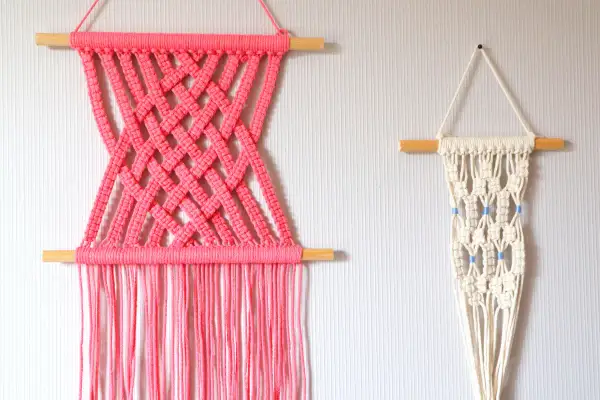 Deux tentures murales en macramé : une rose vif avec un motif croisé, et une écru avec des perles bleues. Idéal pour un tuto macramé mural.