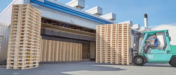 Un chariot élévateur déplace une pile de palettes traitées devant un entrepôt.